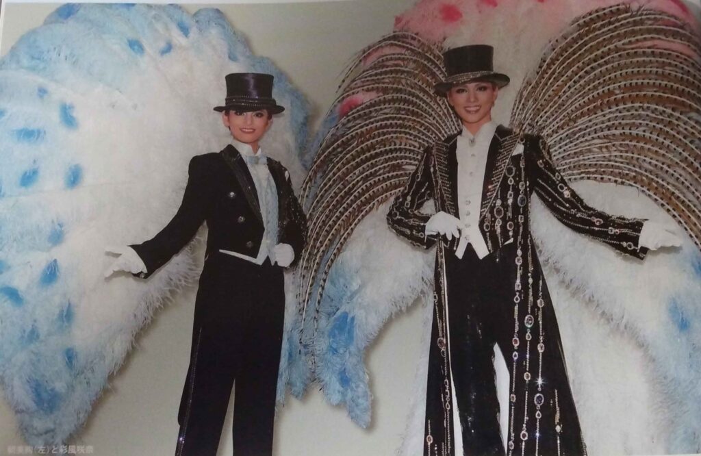 Asami Jun (left) and Ayakaze Sakina (right) posing in their finale outfits for Jewel de Paris. Asami is wearing a black tuxedo with a top hat and white gloves, and wears a large feather pack piece of white feathers with pastel blue tips. Ayakaze wears a black suit with a floor-length coat covered in stripes of metallic chain and large gems. She is wearing a large feather back piece with natural pheasant wing-shaped insets over white feathers with pastel blue and pink tips.