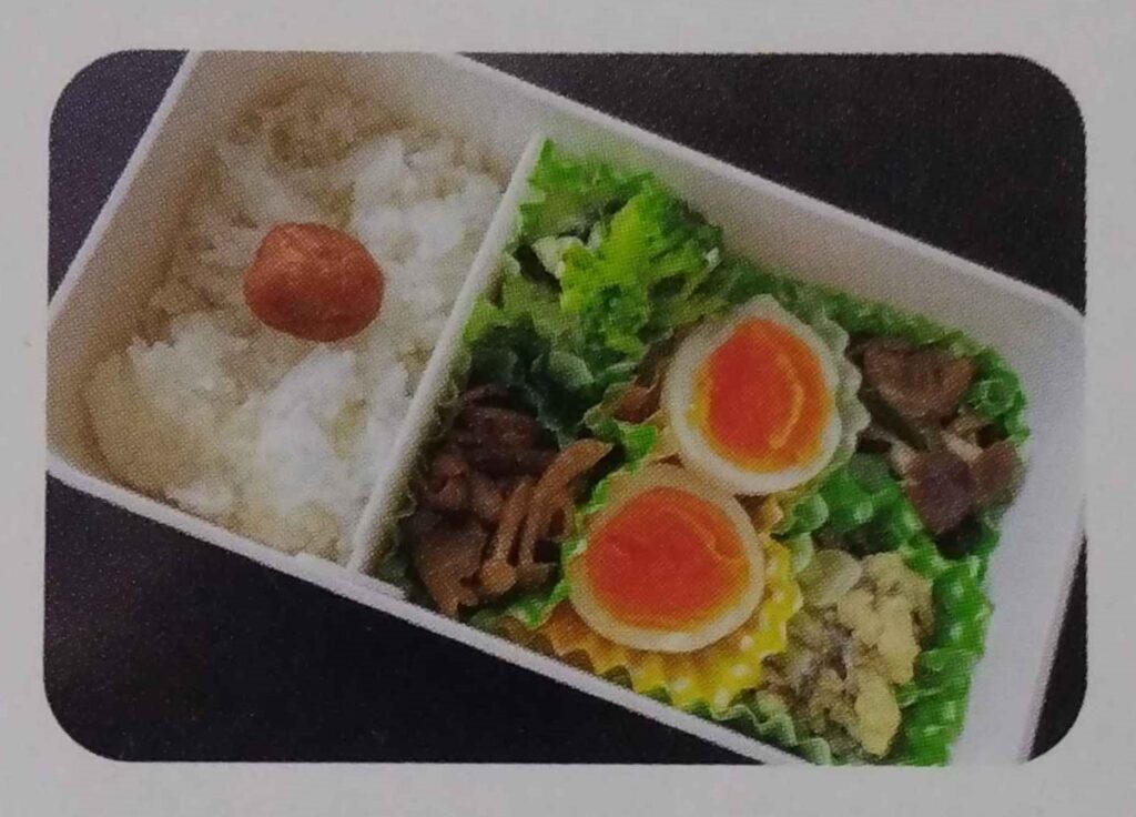 A homemade bento box with a section of rice topped with a pickled plum, and the rest taken up with various other foods including a hard boiled egg.