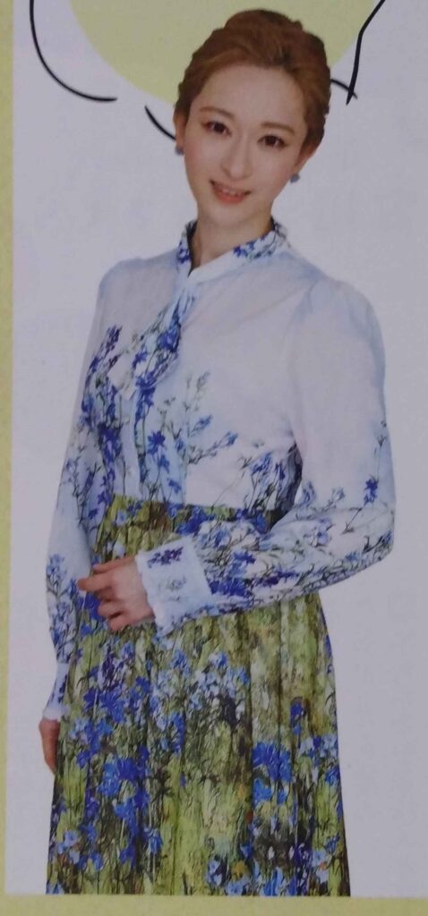 Arisa Hitomi with her hair in an updo. She is wearing a white shirt with soft blue patterning and a green-and-blue skirt in an impressionist absract pattern.