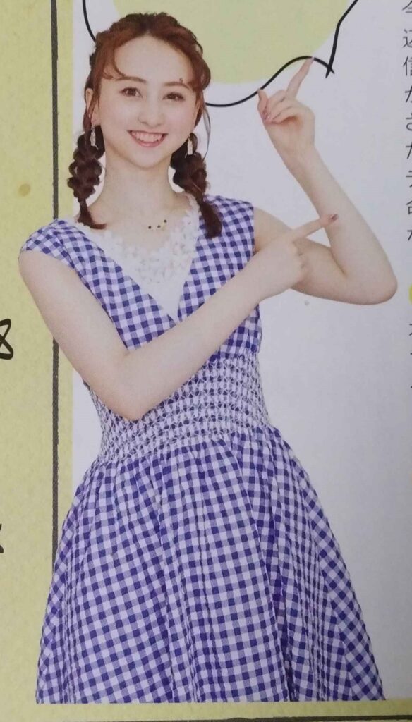 Neiro Yui with her hair in twin fishtail braids, wearing a sleeveless blue-and-white gingham dress with a shirred waist. She is pointing towards the text with both hands.