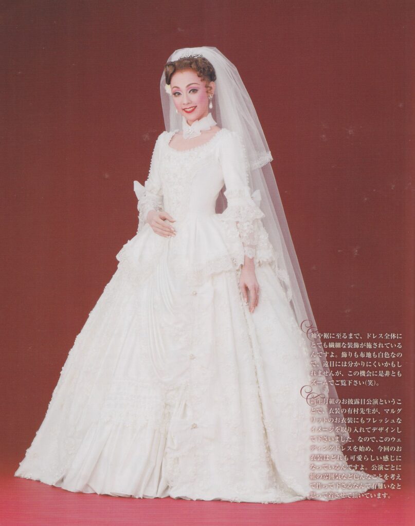 A full-body shot of Aono Yuuki. She wears a large white wedding gown with a full skirt, and wide neckline. She also has a white lace collar-style necklace and a wedding veil that almost reaches the floor.