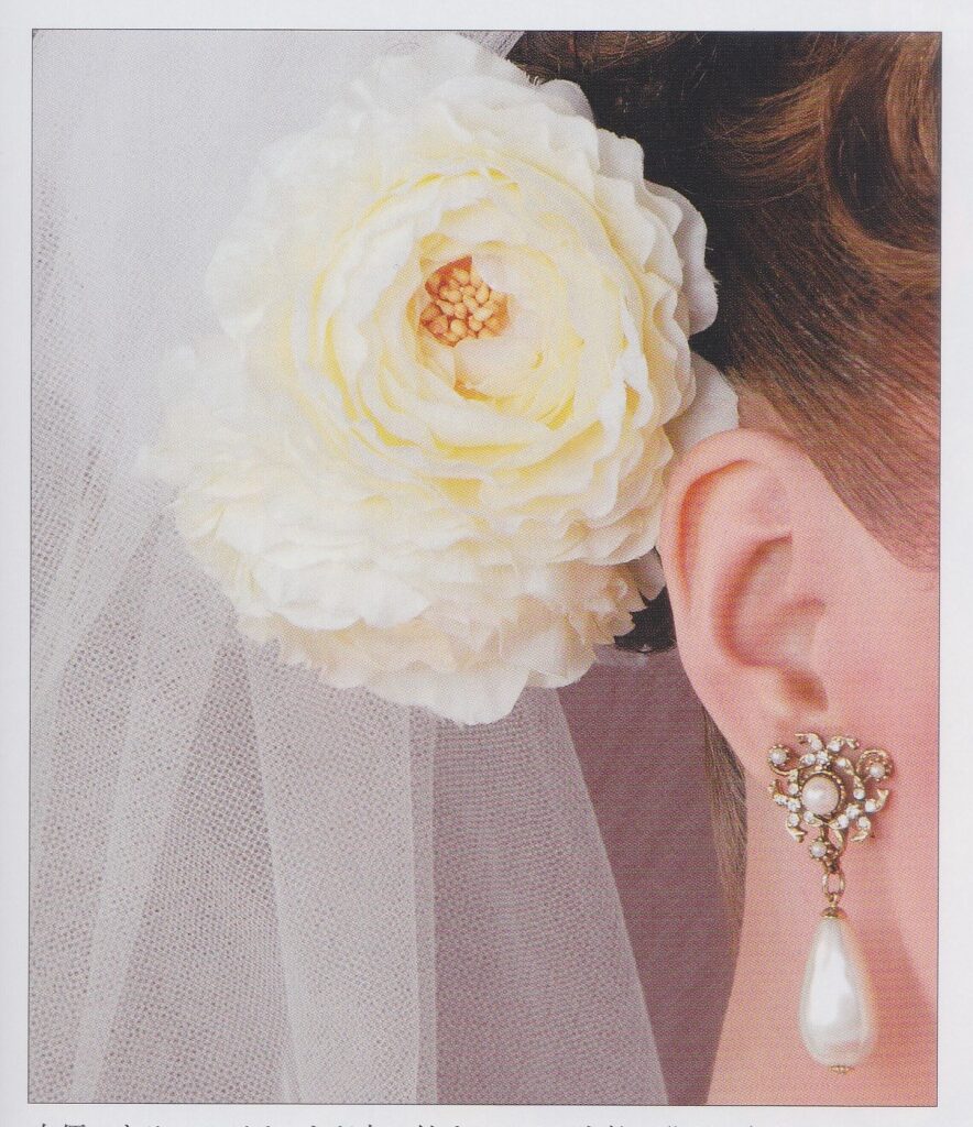 A closeup of Aono Yuuki's hair accessories and earrings. She wears a large off-white fake flower and a pearl drop earring with crystals and a smaller pearl stud.