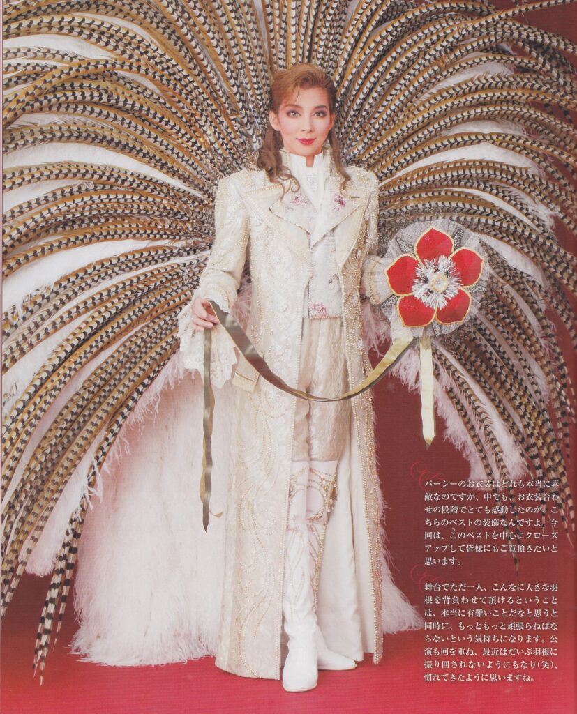 A full-body shot of Kiriya Hiromu in her Scarlet Pimpernel finale costume. She wears a full-length white Regency-styled suit with knee high white boots and a curled auburn wig. She is wearing a wide feather back piece with natural pheasant feathers over white. She holds a shanshan shaped like a pimpernel flower with gold ribbons.