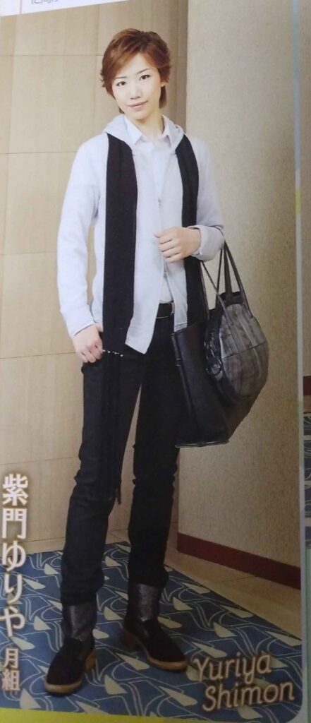 Shimon Yuriya wearing a white shirt and white jacket with black scarf, pants, and ankle boots, holding a black leather tote and smaller silver round bag.