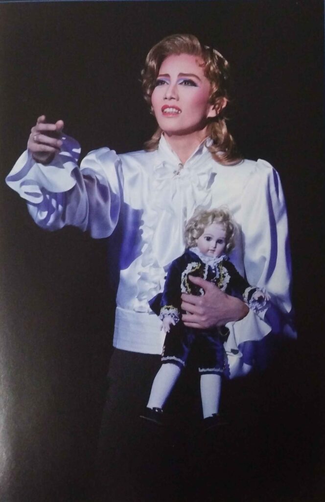 Sou Kazuho with her left hand held out. She is dressed as Fersen, wearing a white ruffled shirt and holding a porcelain doll dressed in a blue suit.