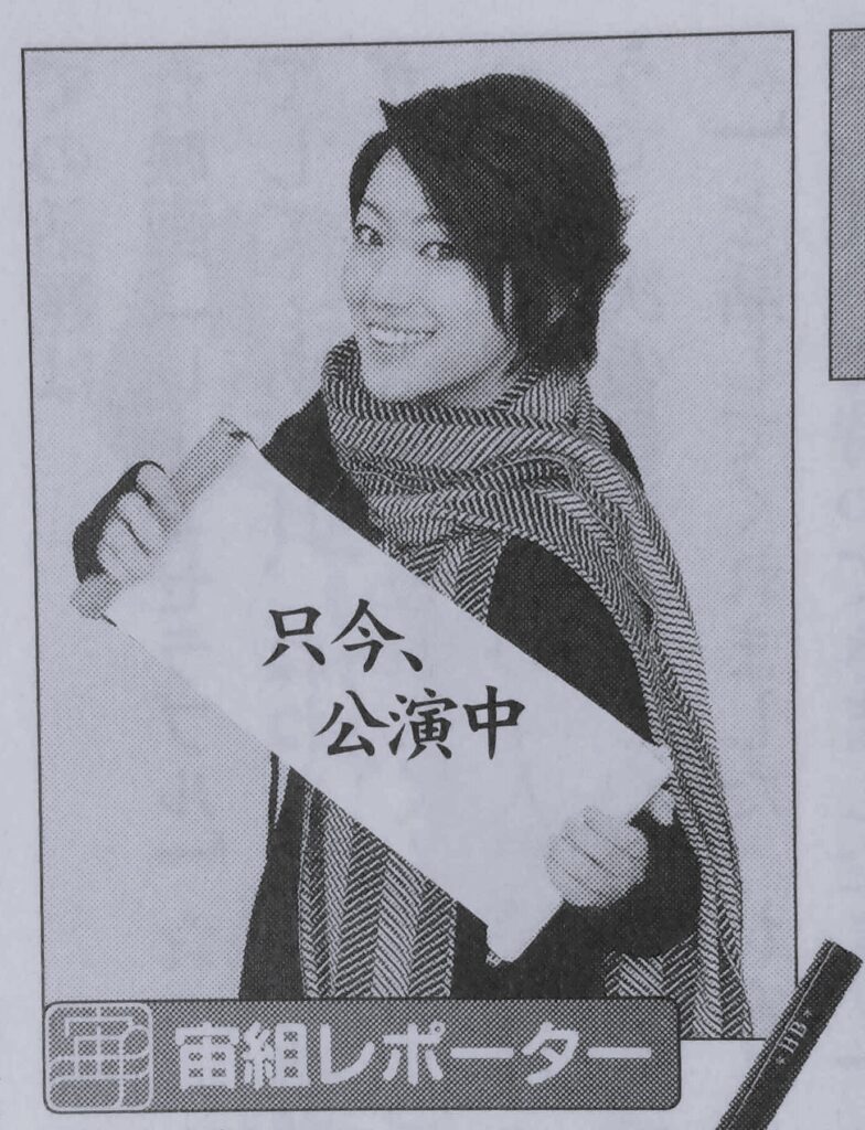 Nanami Hiroki wearing a scarf and fingerless gloves. She is holding up a scroll that has the Japanese text for 'Performing now' written on it.