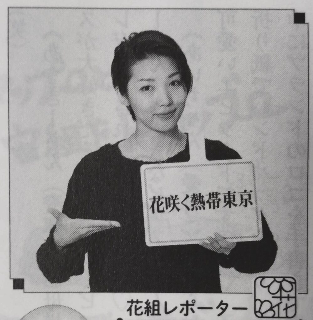 Ootori Mayu wearing a simple dark long-sleeved top. She is holding a sign with the Japanese text for 'Flowery Sweltering Tokyo' with one hand and gesturing towards it with the other hand.