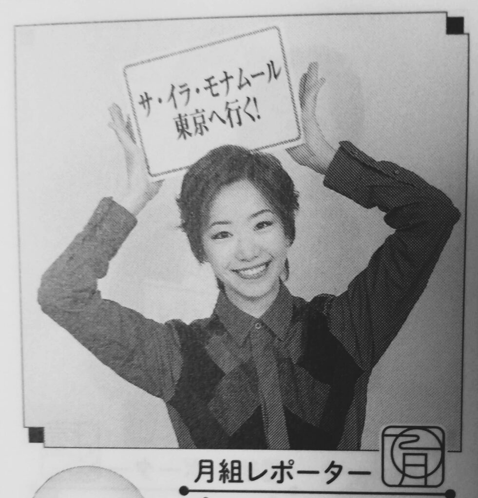 Shimon Yuriya wearing a long sleeved shirt with checkered front panels. She is smiling and holding a sign on her head with the Japanese text for 'Ca Ira Mon Amour to Tokyo'.