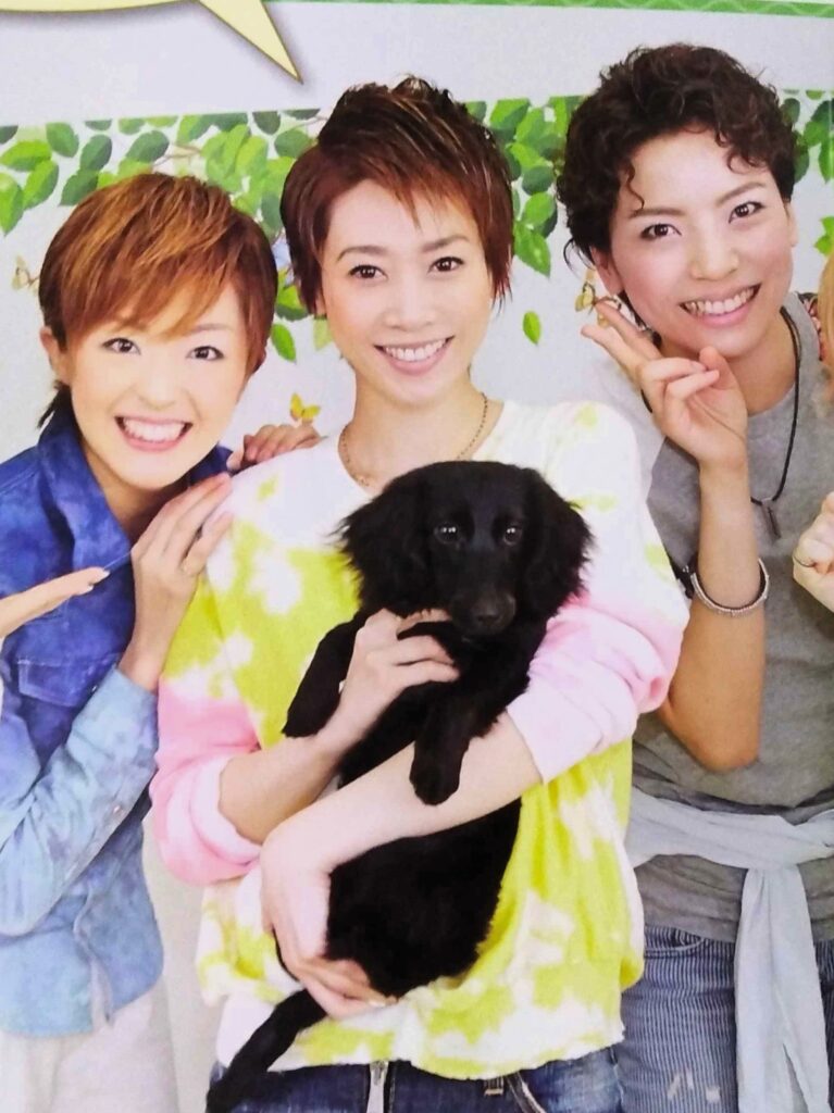 Masaki Ryuu holding a black dachshund puppy. The photo is cropped to show Kizuki Yuuma and Akizuki Saya next to her.